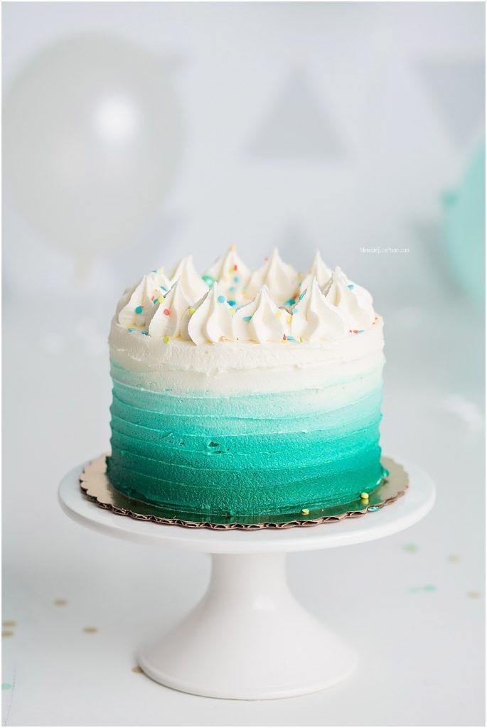 Beautiful ombre teal cake with white frosting peaks displayed on white cake stand against light background.