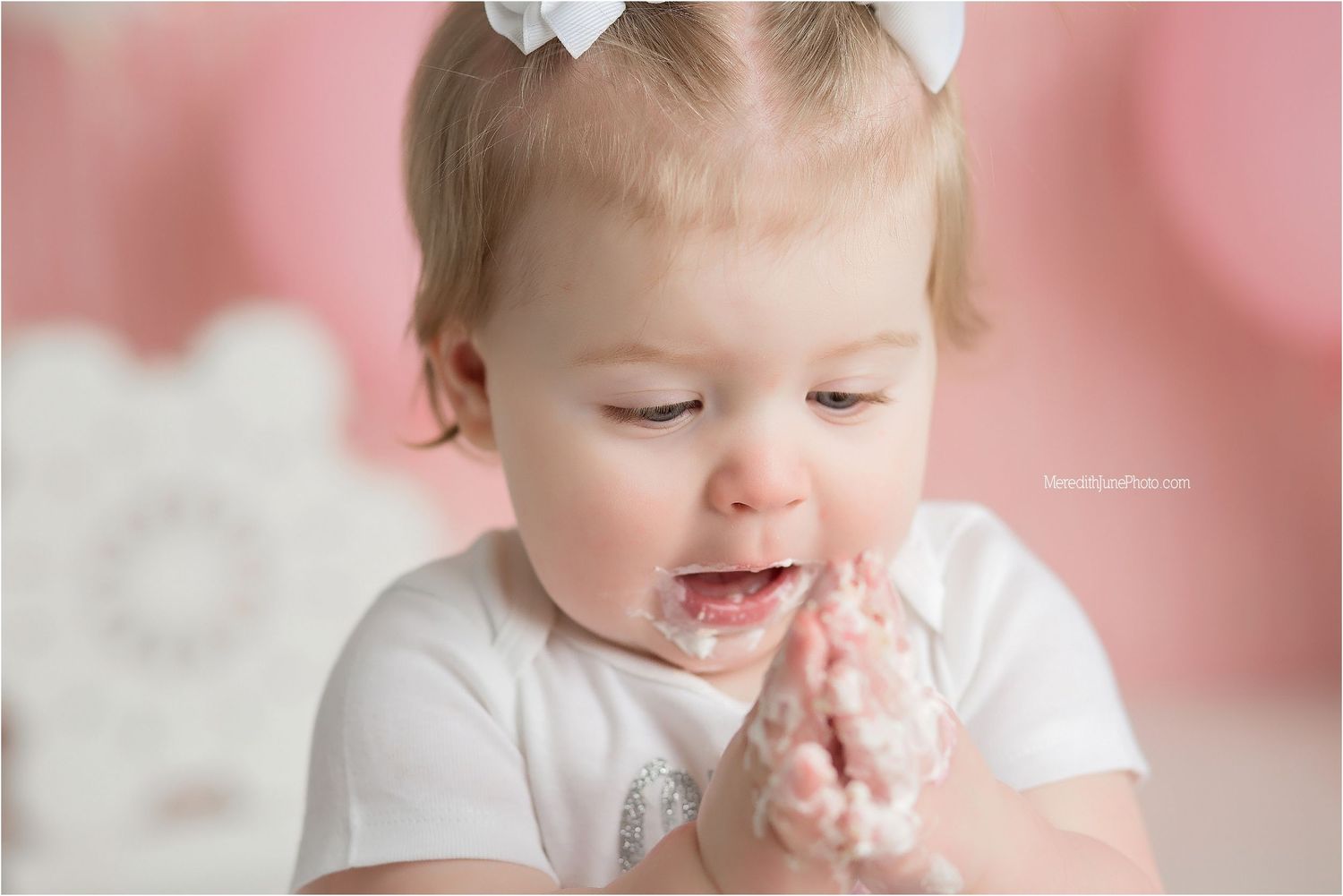 A baby enjoying cake during a first birthday photoshoot with pink decorations and white lace accents.