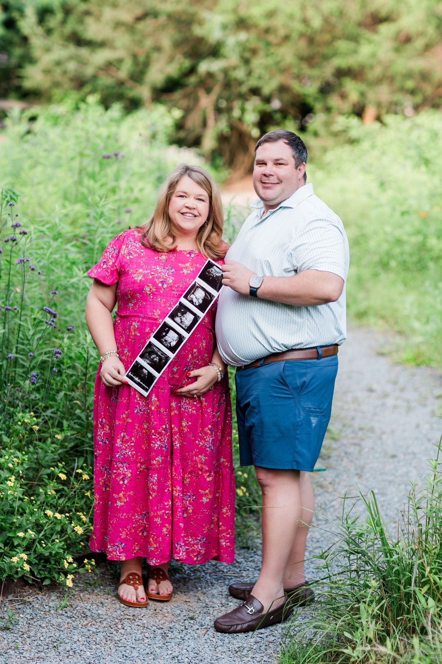 A couple in pink dress and casual wear pose together on a gravel path surrounded by greenery during an outdoor photoshoot.