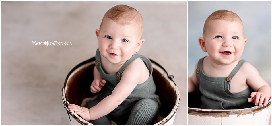 A smiling baby sits in a dark wooden bowl wearing an olive green sleeveless outfit against a light backdrop.