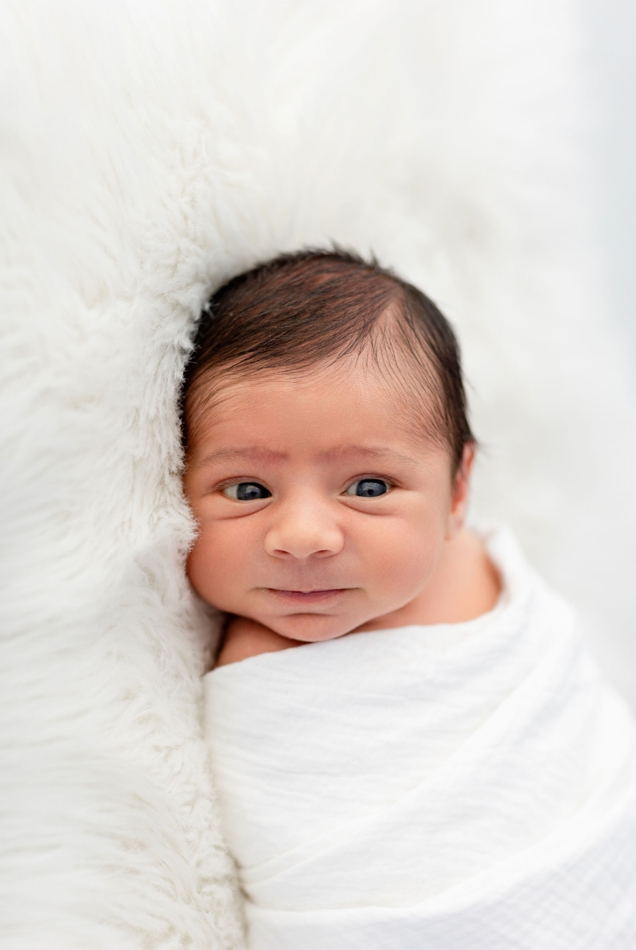 A newborn baby swaddled in white blankets on a soft white background.