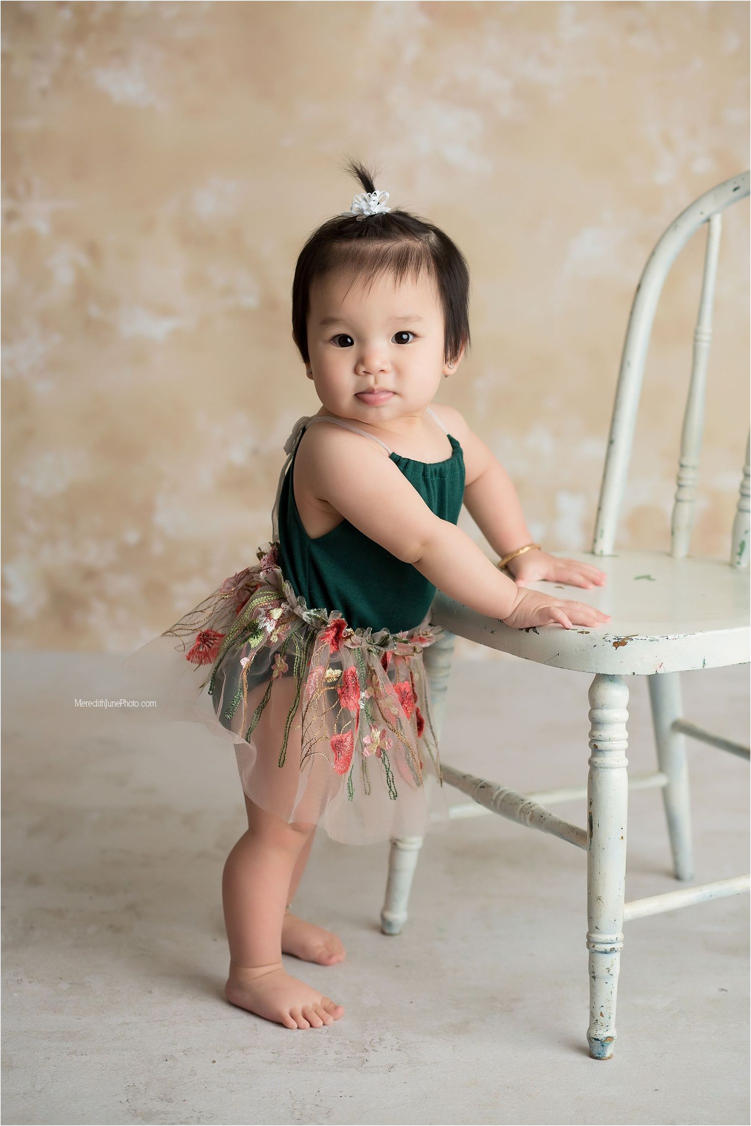 A small child in a green and floral tutu dress plays near a vintage white wooden chair in a studio setting.