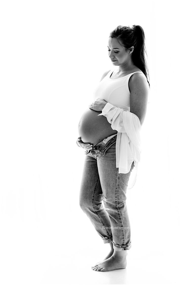 Black and white maternity photo of an expectant mother in jeans and tank top standing barefoot against white background.