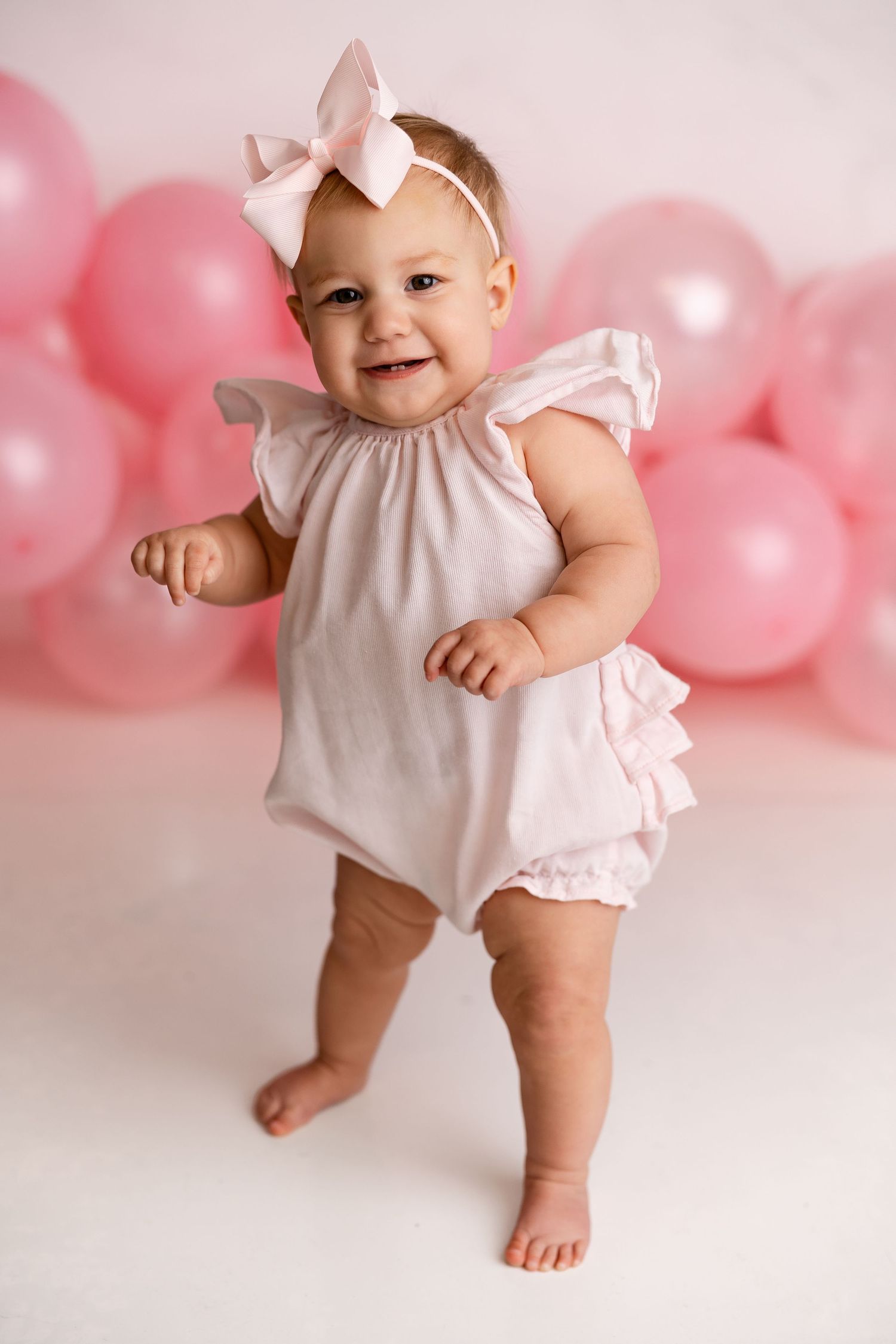 Sweet baby in white ruffled romper and bow headband poses against pink balloon backdrop for milestone photo.