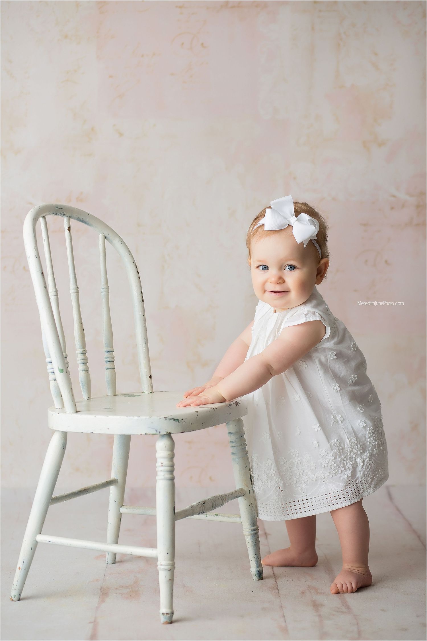 A baby in a white dress interacts with a vintage white wooden chair against a neutral backdrop.
