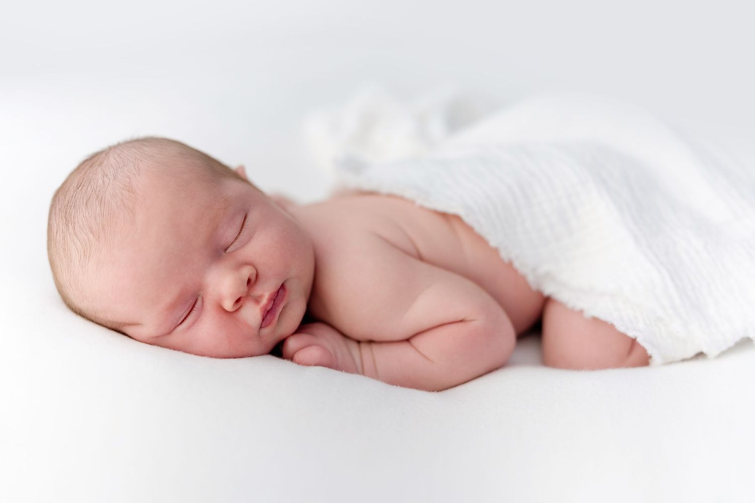 Peaceful newborn baby sleeping wrapped in a soft white blanket on a white background.