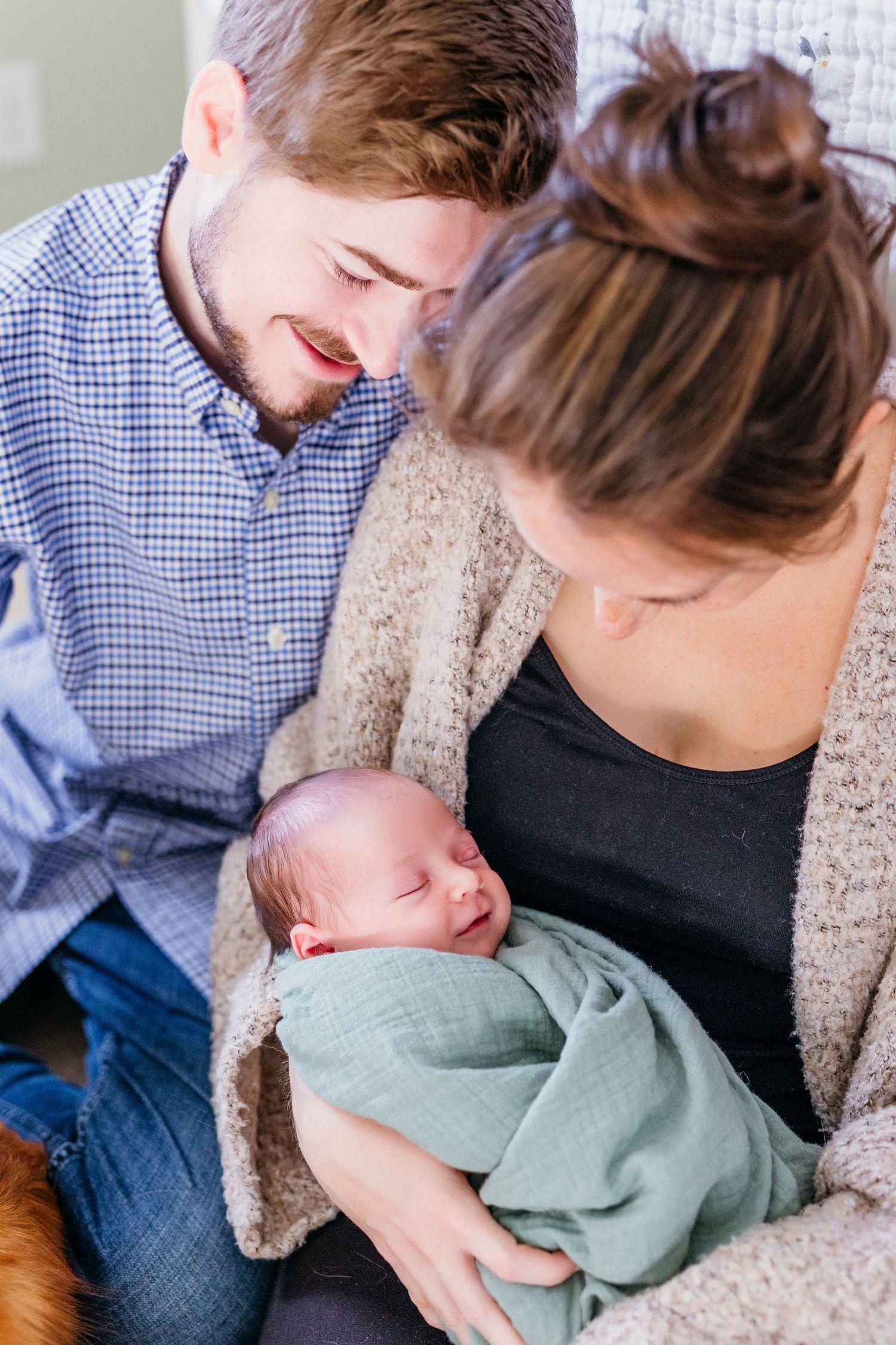 A couple tenderly holds their sleeping newborn baby while sitting together on a couch.