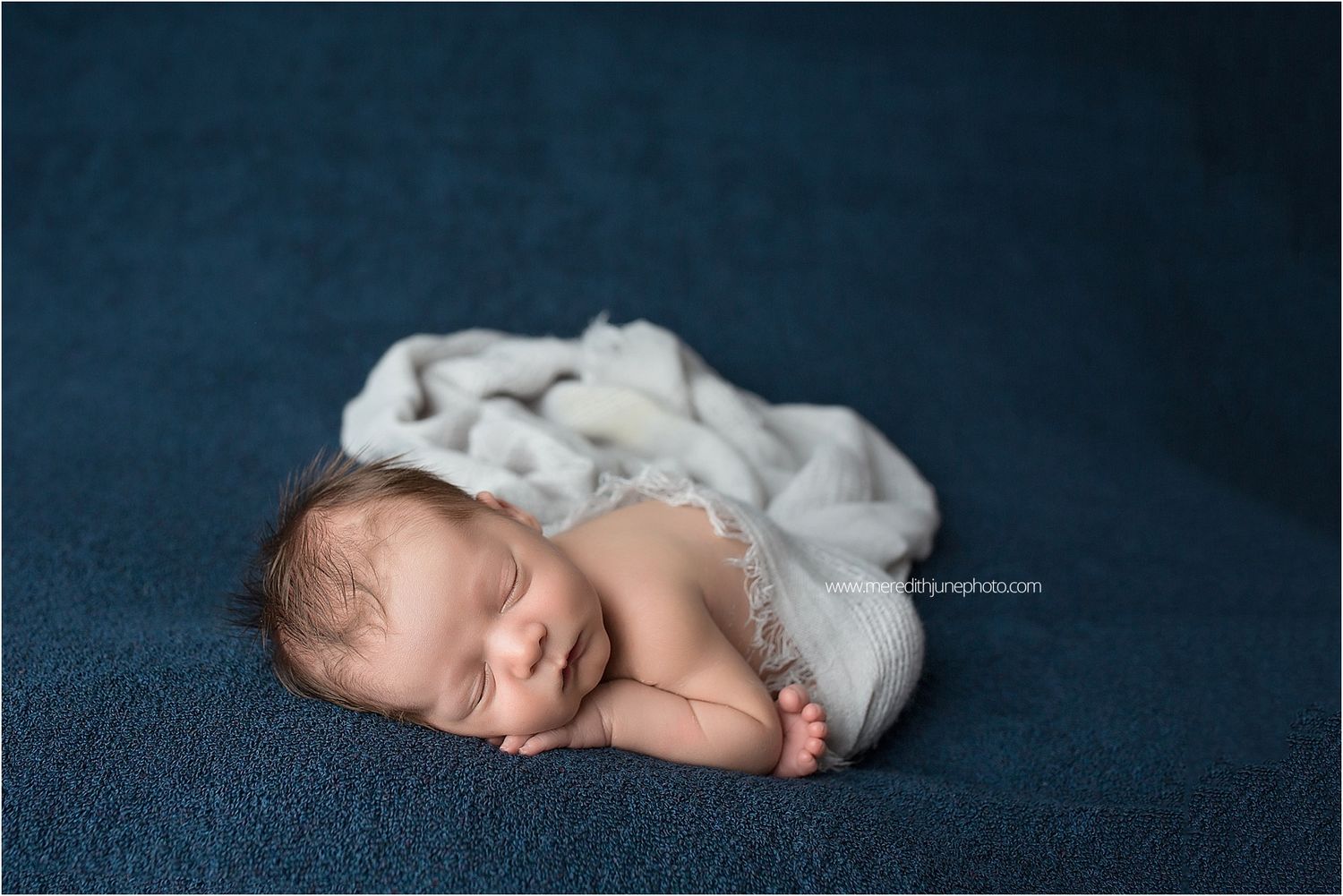 Peaceful newborn baby wrapped in soft white blanket sleeping on dark blue backdrop during professional photo session.