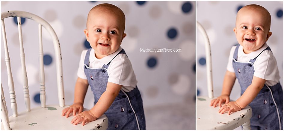 A baby in blue and white overalls smiles while leaning on a white chair against a polka dot background.