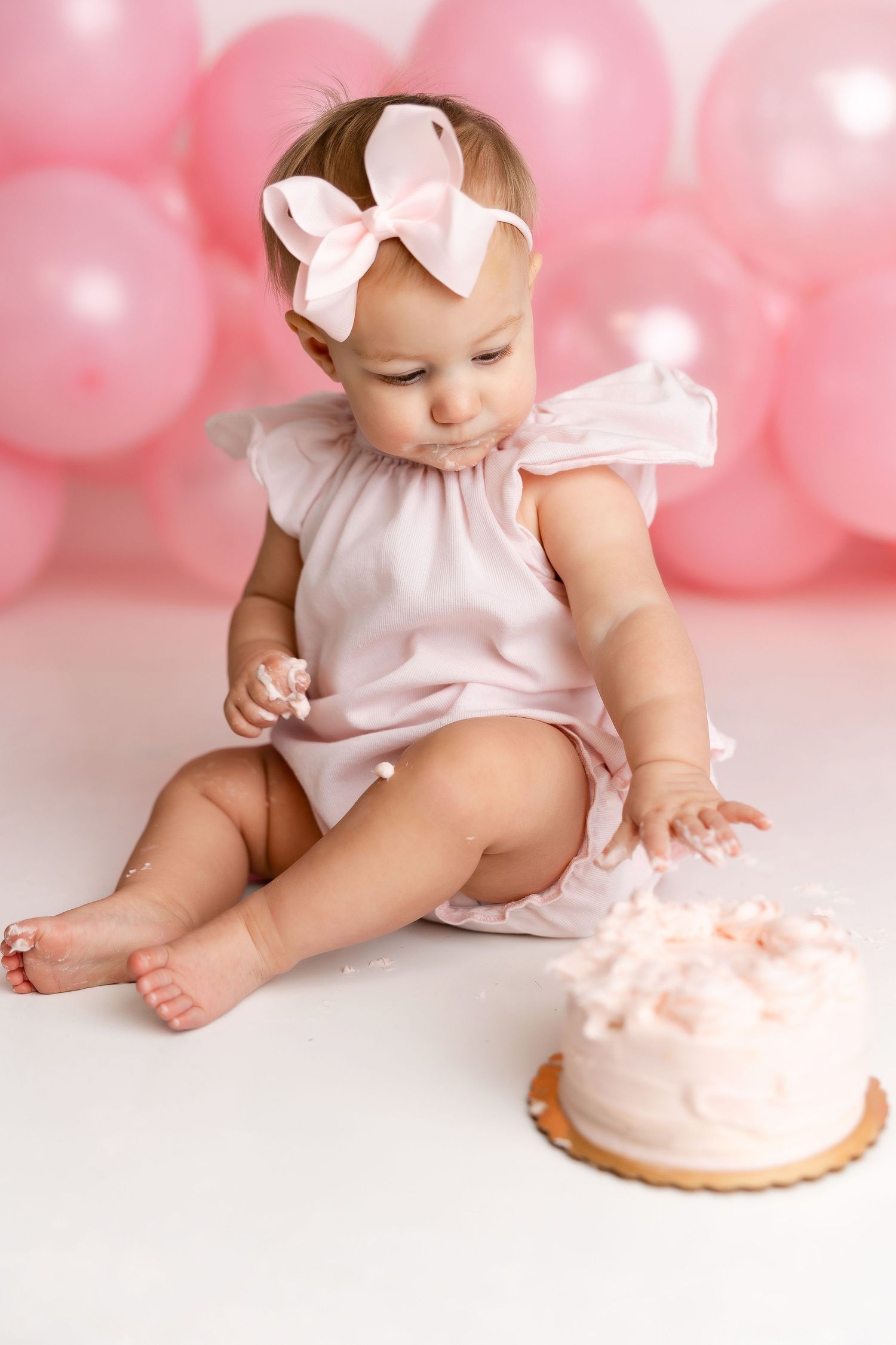 Baby in pink dress and bow sitting beside pink birthday cake with pink balloon backdrop for first birthday photoshoot.