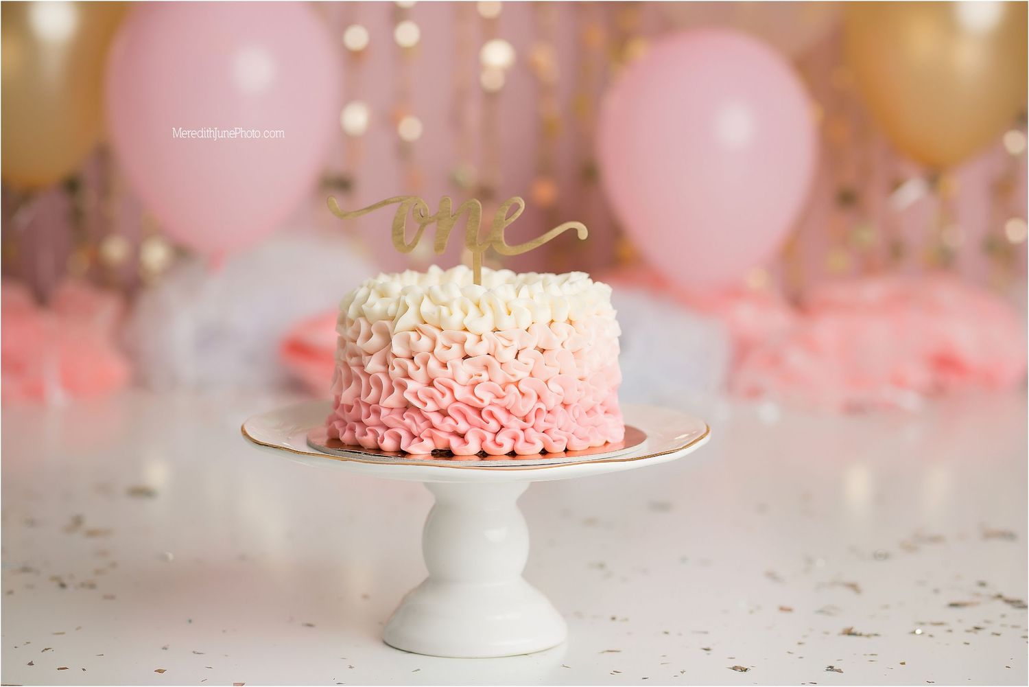 Pink ruffle cake on white pedestal stand with blurred bokeh lights and pink balloons in background.