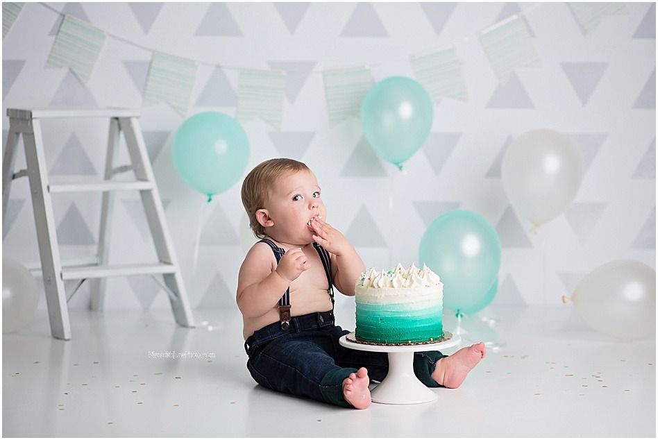A baby in navy blue shorts poses with mint green balloons and a cake for a birthday photoshoot.