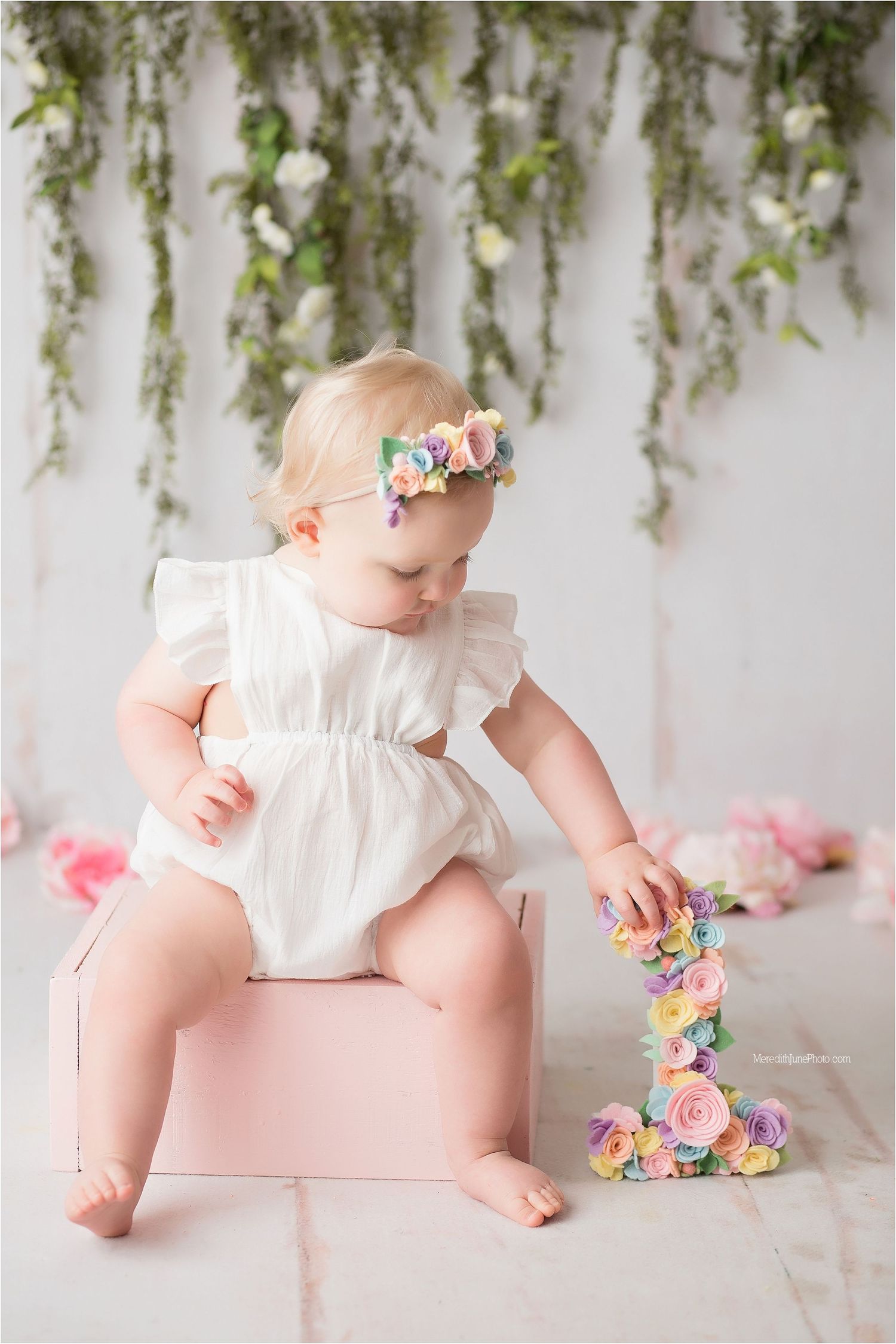 Baby in white dress sits on pink backdrop with colorful floral decorations in professional studio setting.