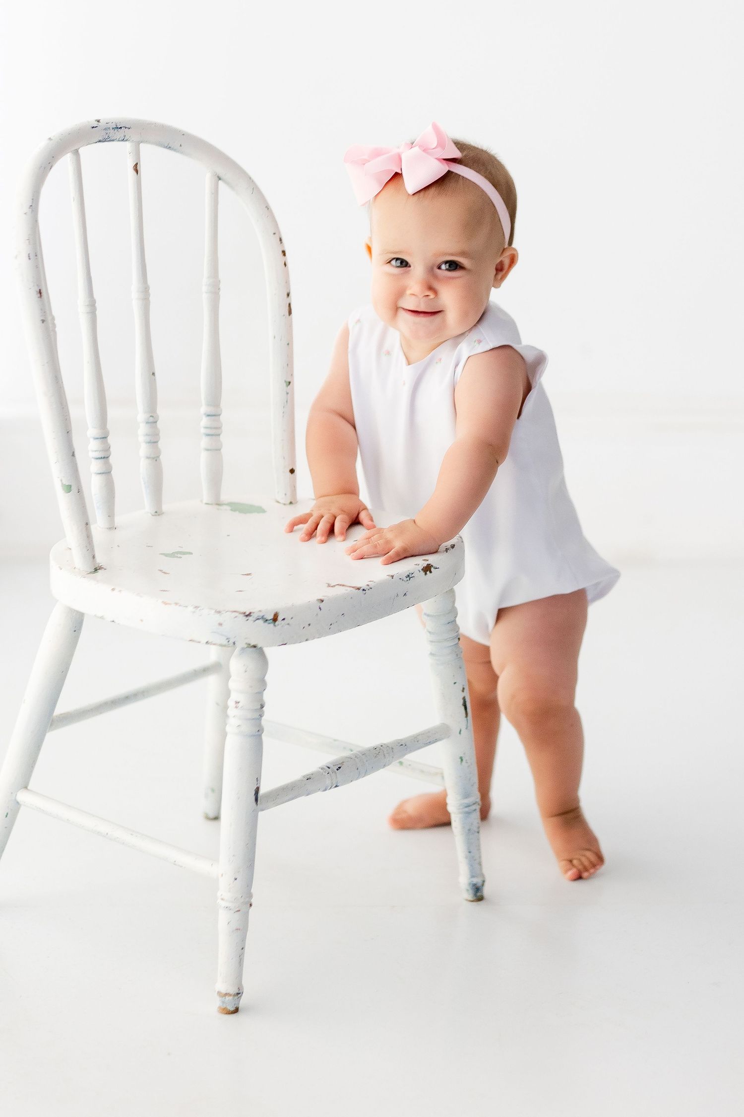 Baby in white outfit holds onto distressed white wooden chair while standing on white background.