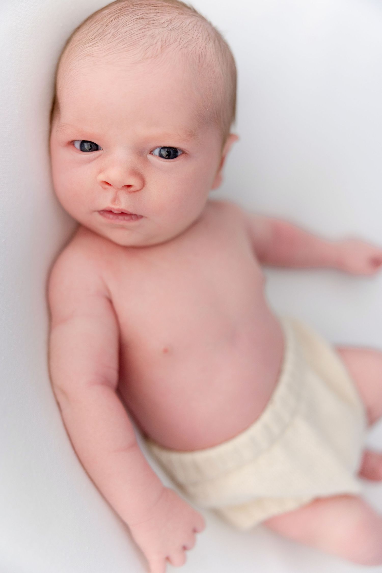 Newborn baby with blue eyes lying on white background wearing a cream diaper, looking up at the camera.