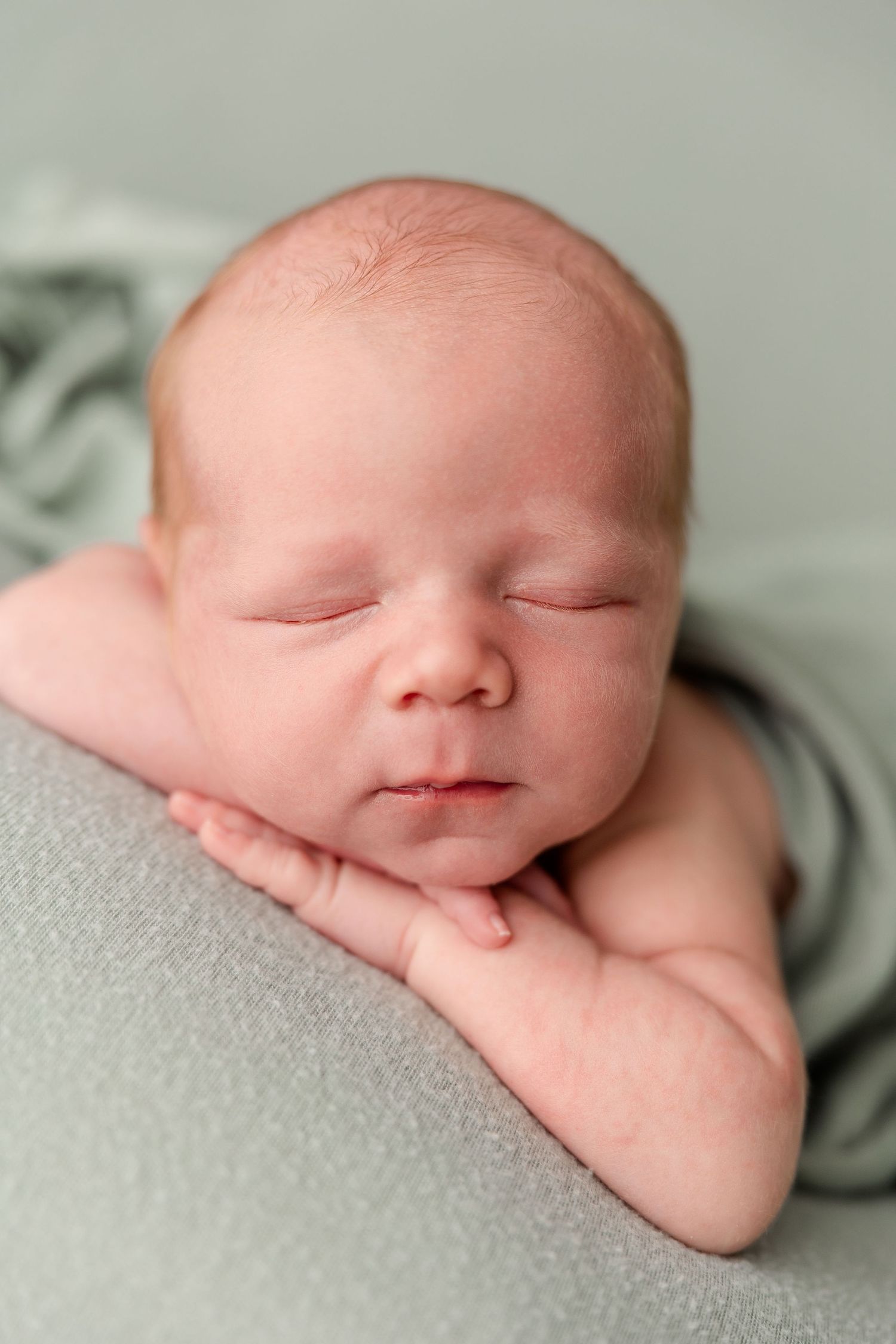 Peaceful newborn baby sleeping with hands tucked under chin on soft sage green fabric backdrop.