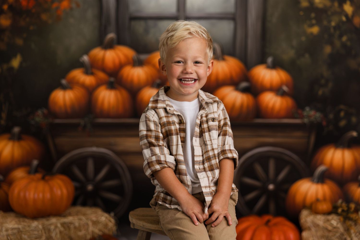 A cheerful young boy smiles in front of a festive autumn backdrop with orange pumpkins and a wooden cart.