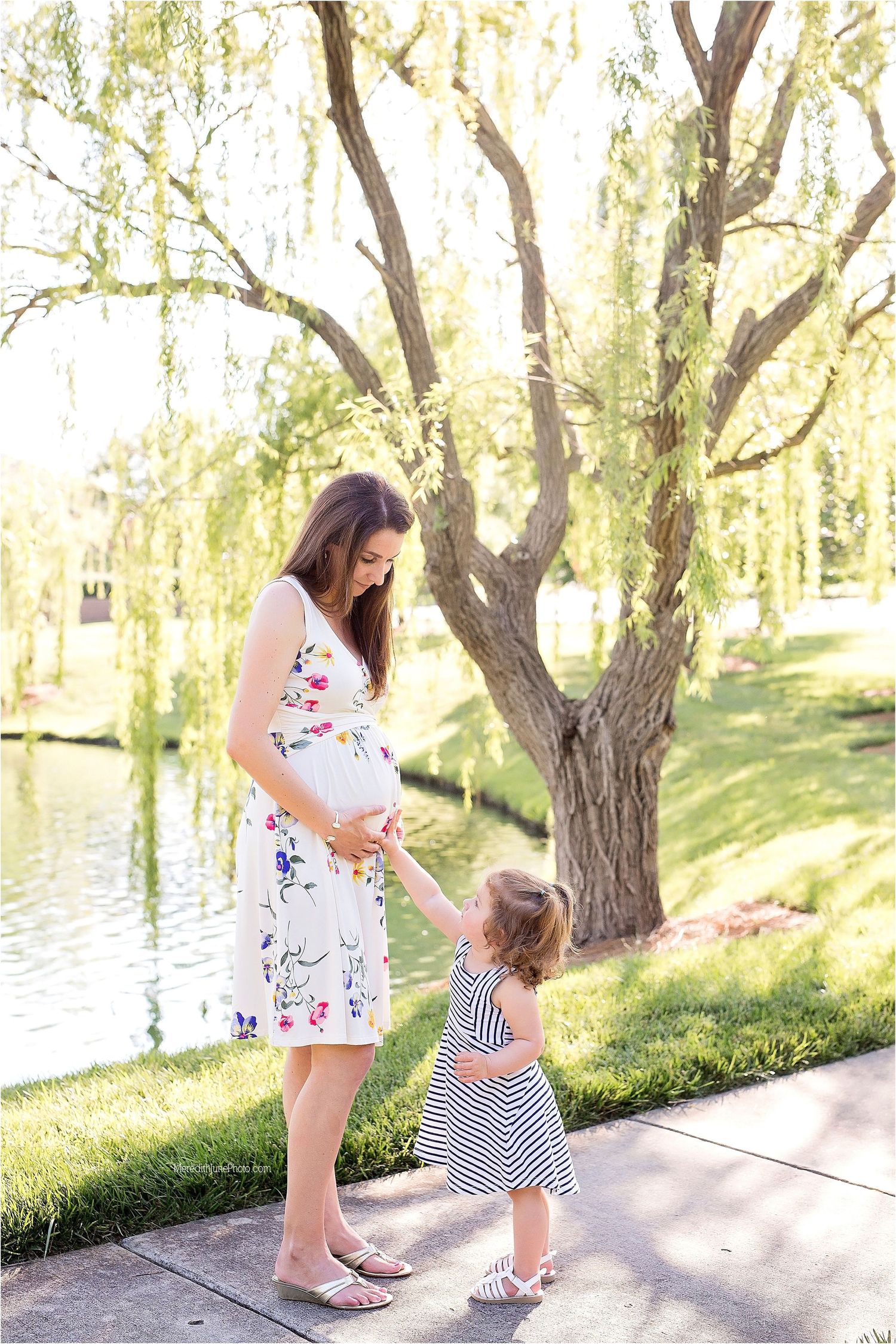 A mother and child in floral dresses walk together along a scenic riverside path on a sunny spring day.