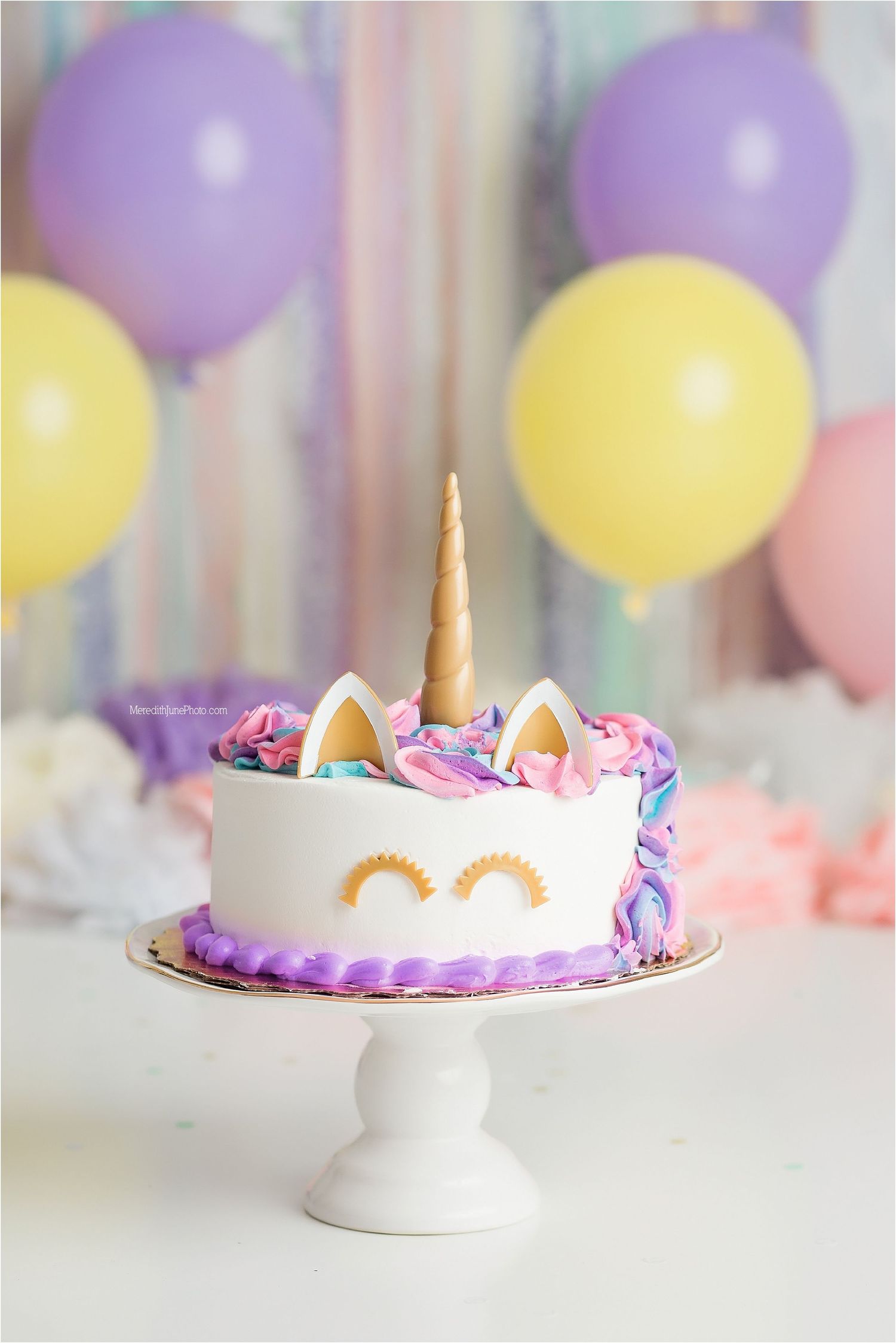 A unicorn themed birthday cake with pink and purple frosting on a white pedestal stand with pastel balloons in background.