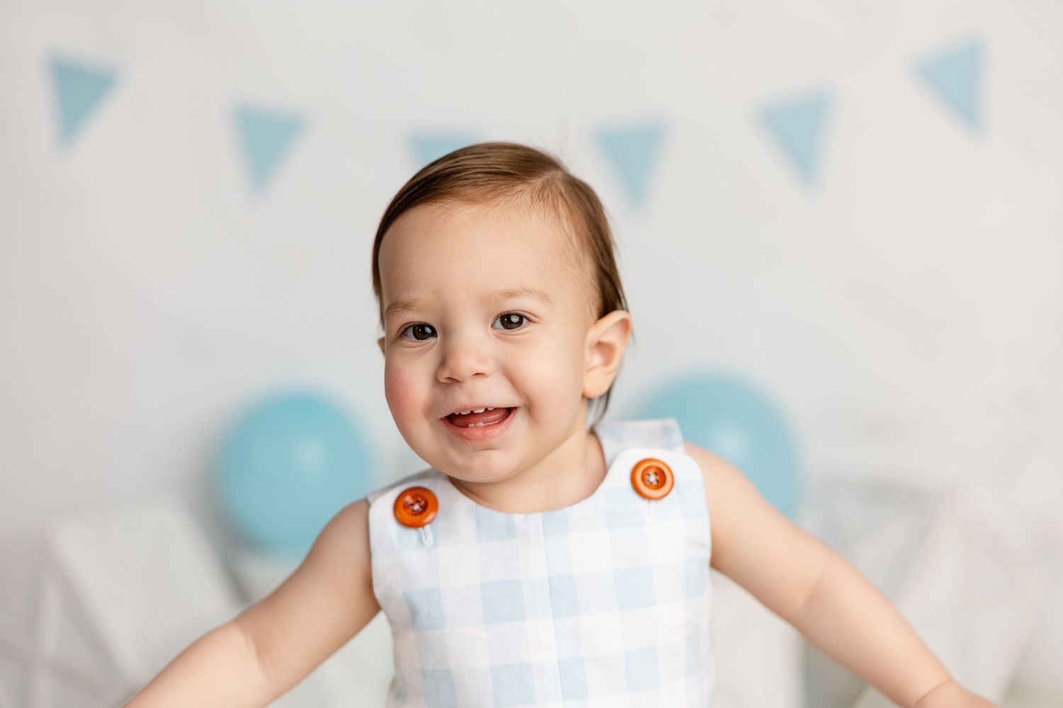 A baby in a white dress smiles while sitting against a blue and white bunting banner background.