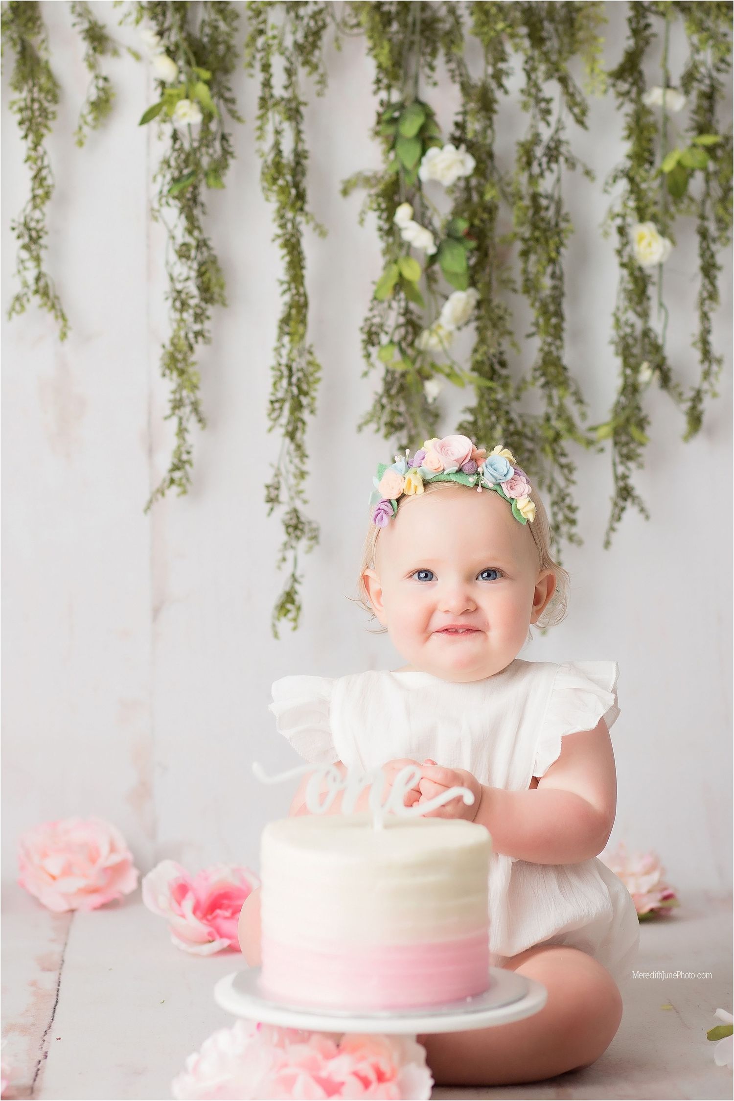 Cake smash photo session featuring white and pink birthday cake with greenery backdrop and delicate floral decorations.