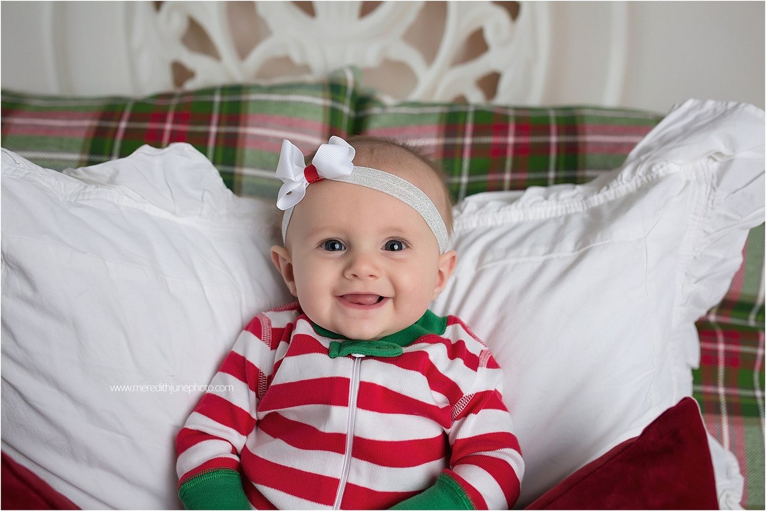 Small child in red and white striped holiday outfit smiling while sitting on plaid bedding with decorative backdrop.