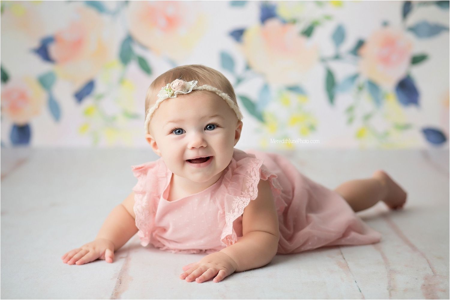 Baby in pink ruffled dress lying on floor against floral backdrop during professional photo session.