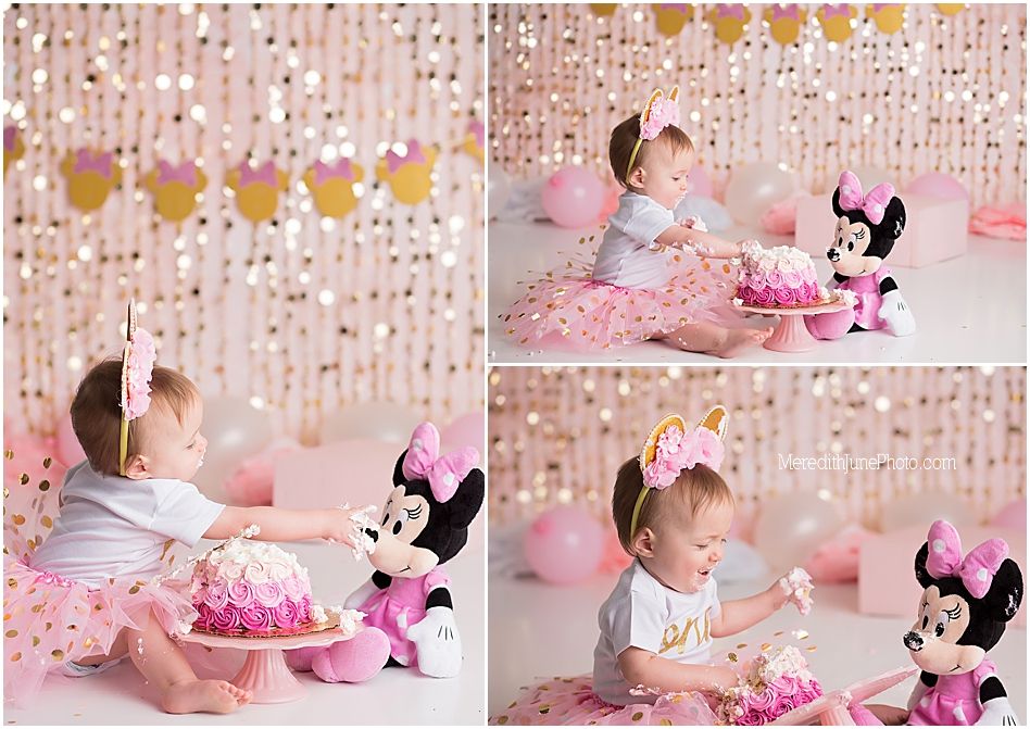 Baby in pink dress celebrates first birthday with Minnie Mouse plush and cake against sparkly backdrop.