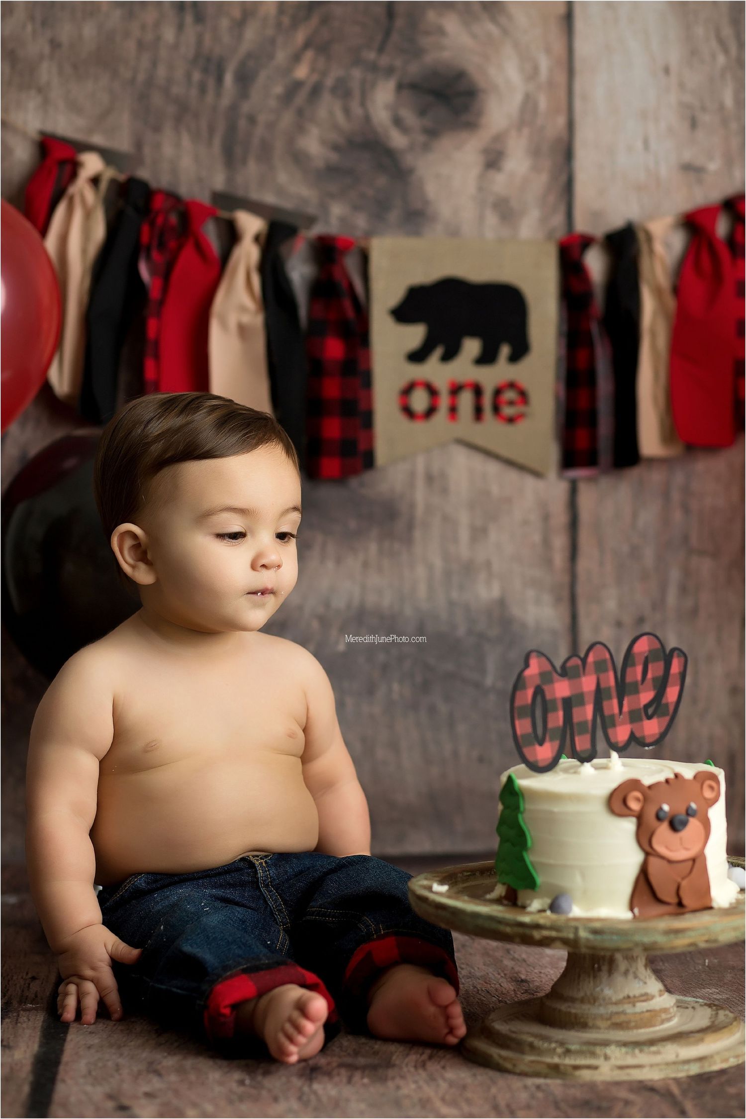 A cake smash photo session featuring buffalo plaid decor and a rustic backdrop with a simple white cake.