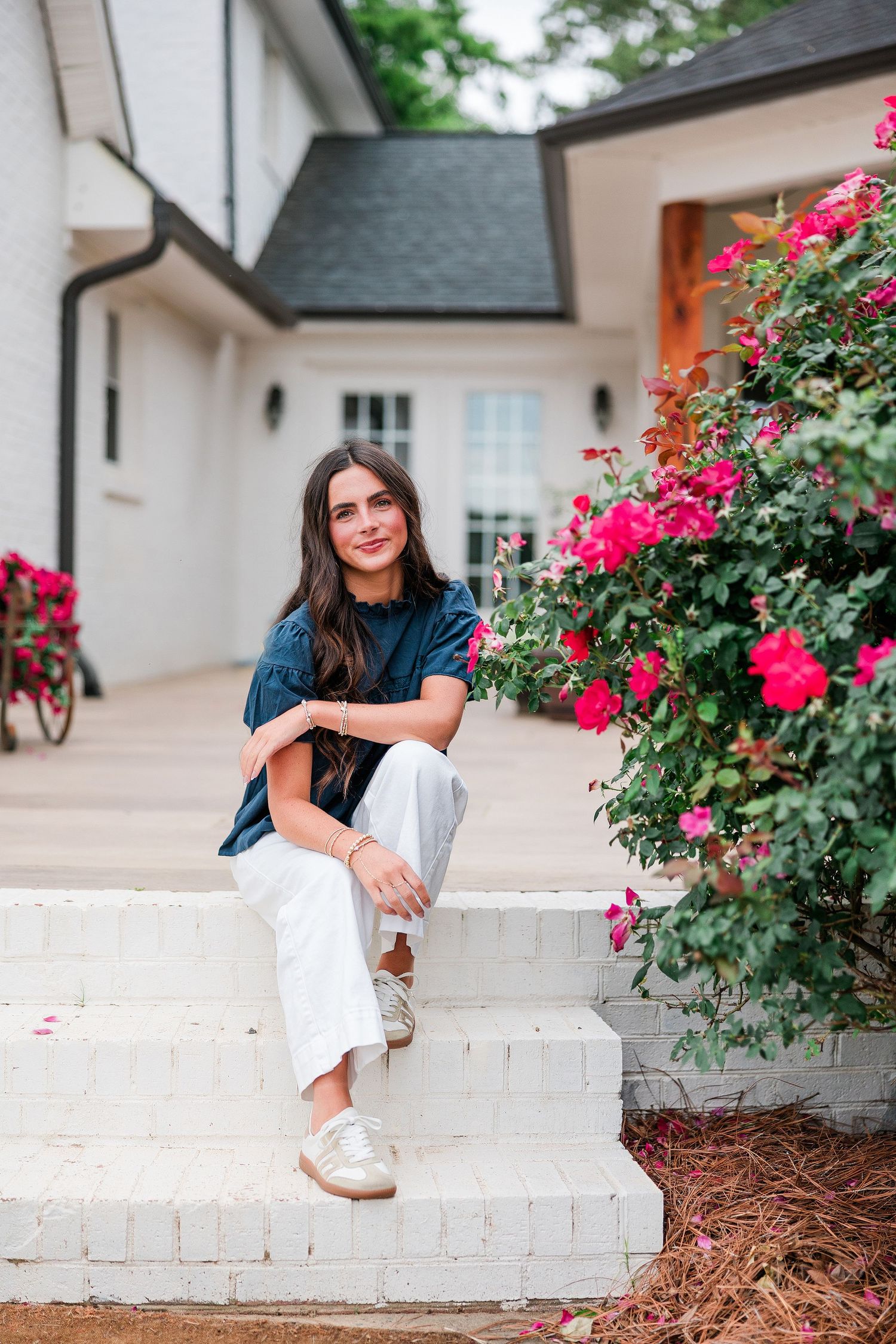 Young woman in navy top and white pants sitting on white brick steps beside vibrant pink rose bushes near a home.