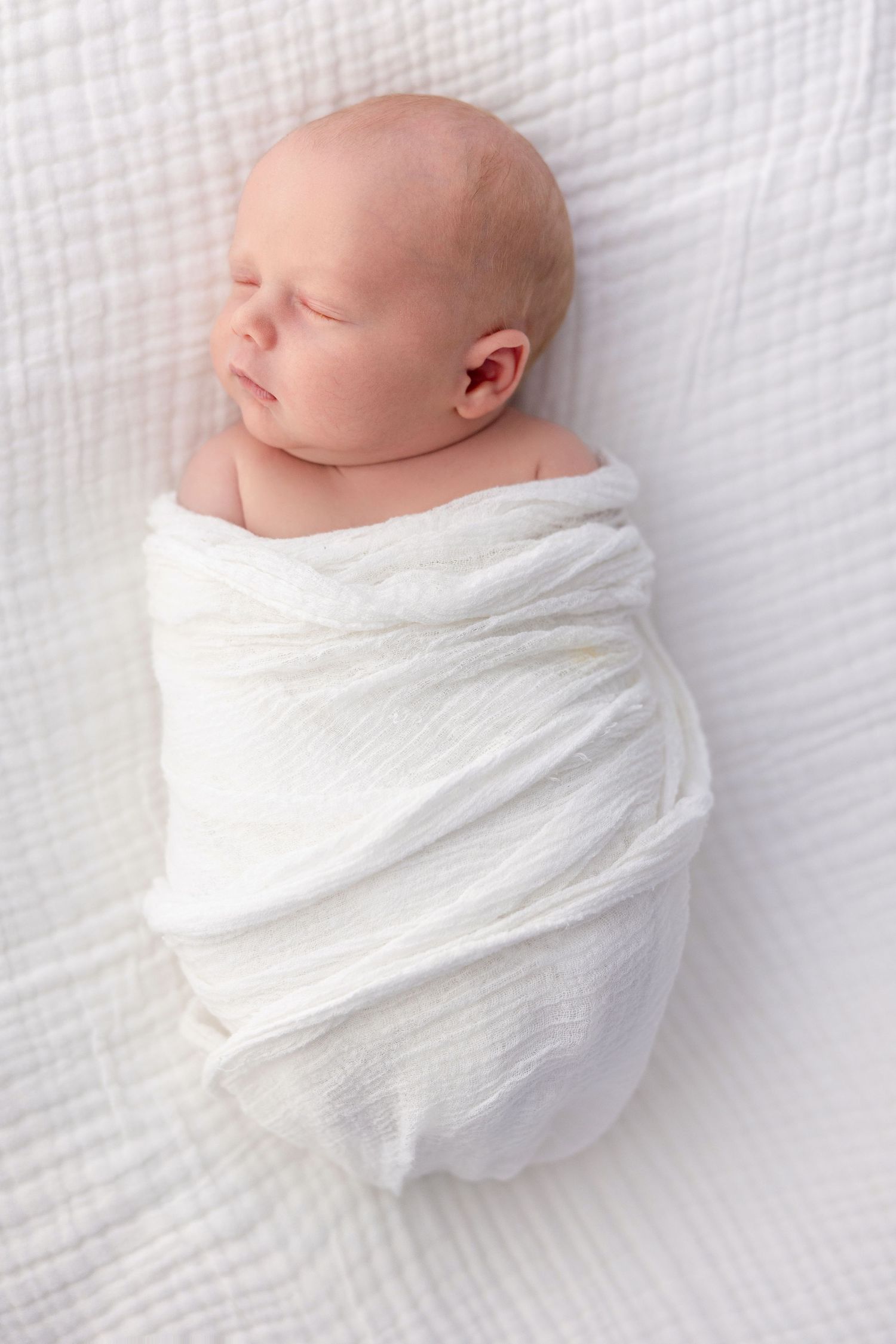 Peaceful newborn baby sleeping soundly while swaddled in a soft white wrap on a white textured blanket.