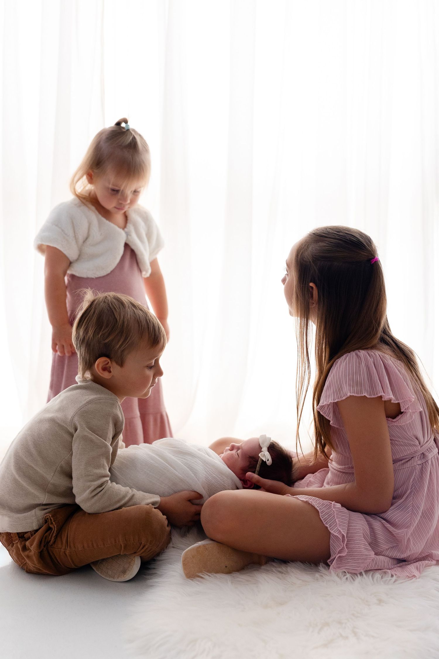 Three young siblings in soft neutral and pink outfits gather around a sleeping newborn baby on a fluffy white rug in a bright studio.