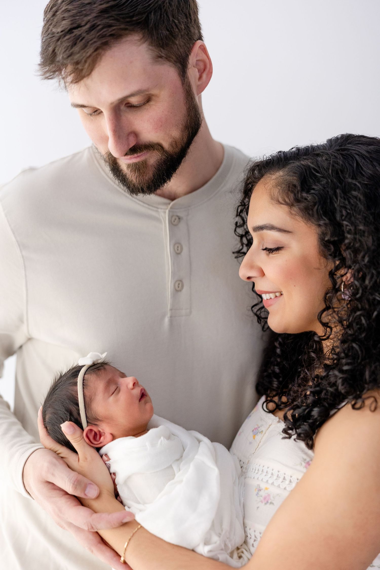 A loving family moment with parents gazing adoringly at their newborn baby in a white onesie against a light background.