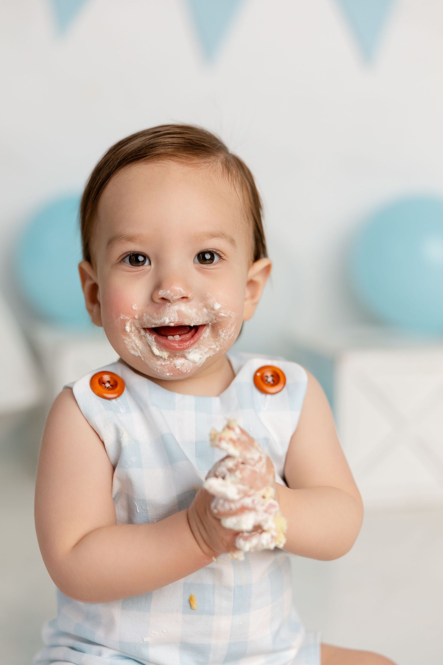 Baby with cake frosting on face smiles during first birthday photoshoot with blue decorations.