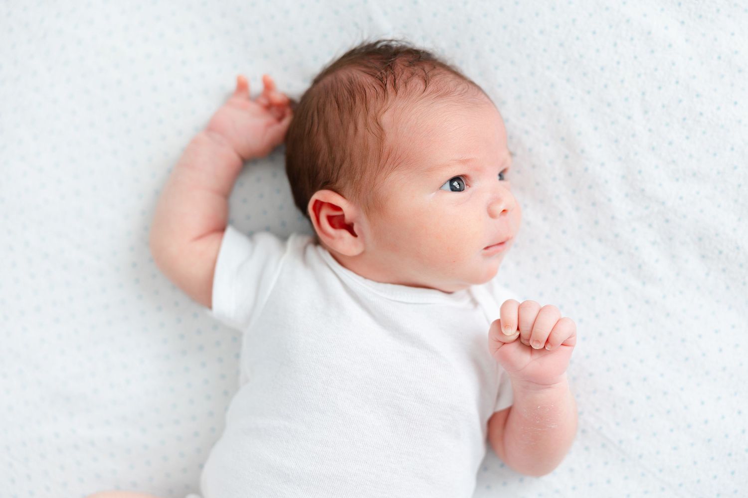 A newborn baby wearing white clothing lies on white bedding while looking to the side.