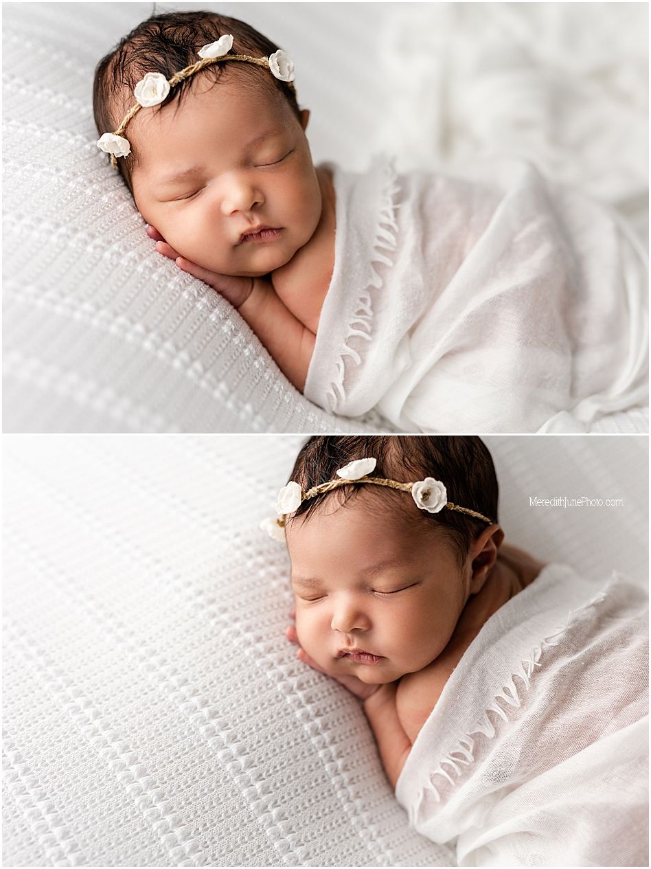 Sleeping newborn wrapped in soft white blanket with delicate floral headband rests peacefully on textured backdrop.