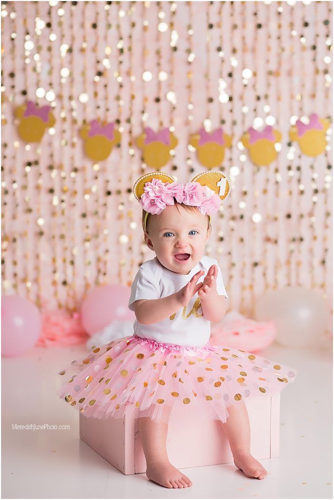 Baby in pink tutu dress and crown sits on floor with sparkly backdrop for first birthday cake smash photo session.