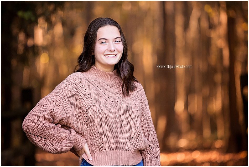 A person in a cozy pink sweater poses outdoors during autumn with warm sunlight filtering through trees.