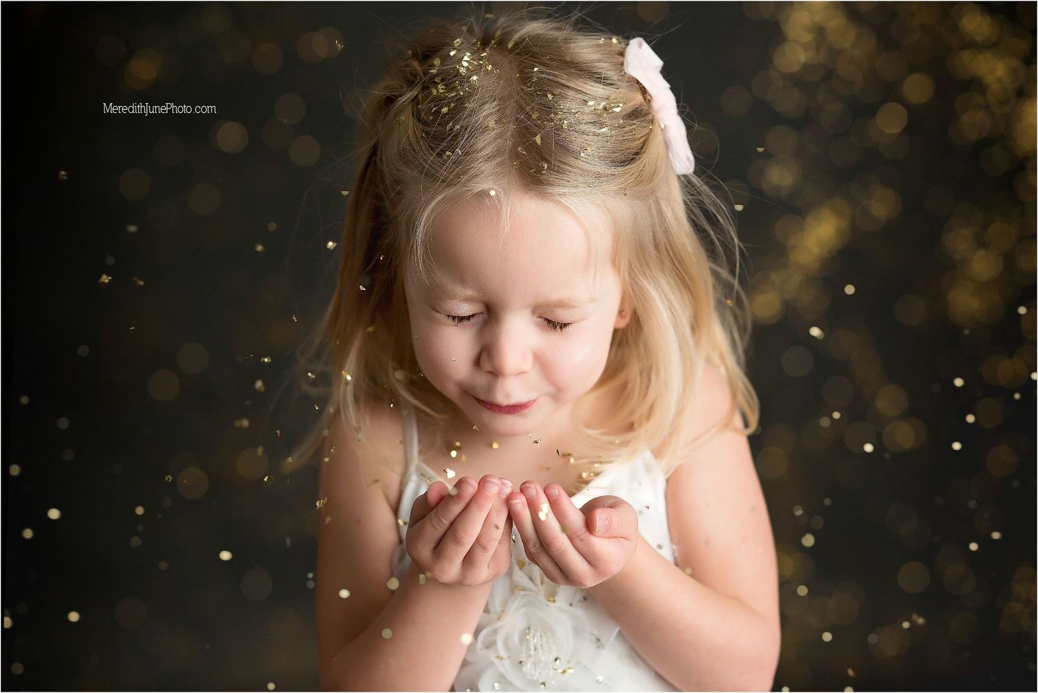 Child in white dress blowing glitter in magical holiday photoshoot against dark background.