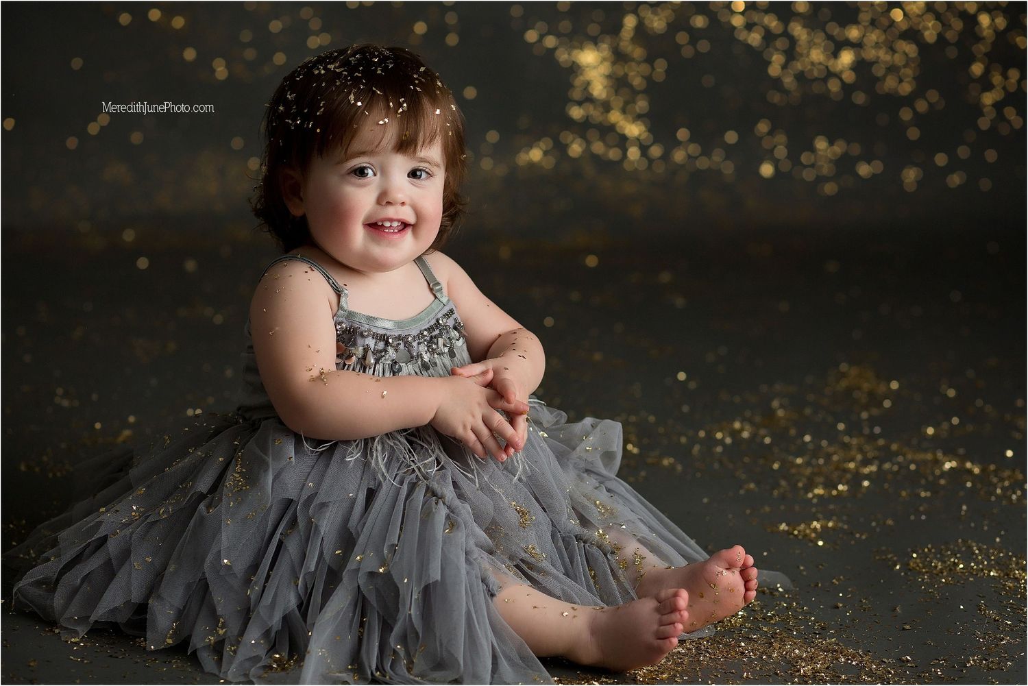 Child in a grey tulle dress sits against a dark backdrop with floating gold glitter.