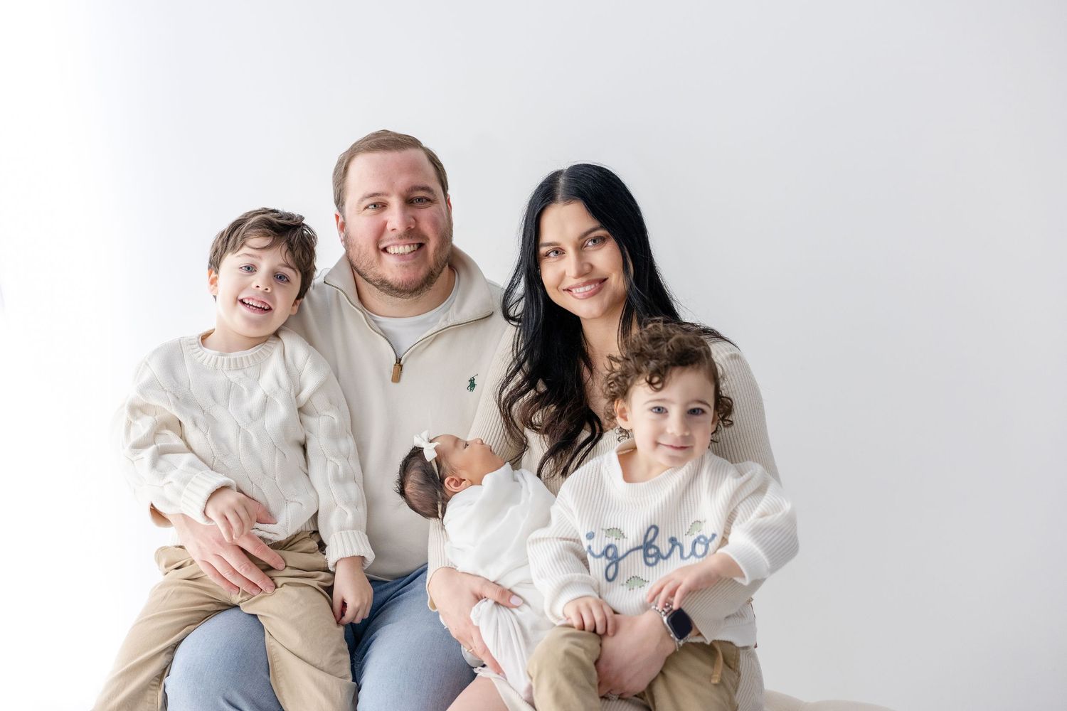 A family wearing coordinated white outfits poses together on a light gray background for a portrait session.