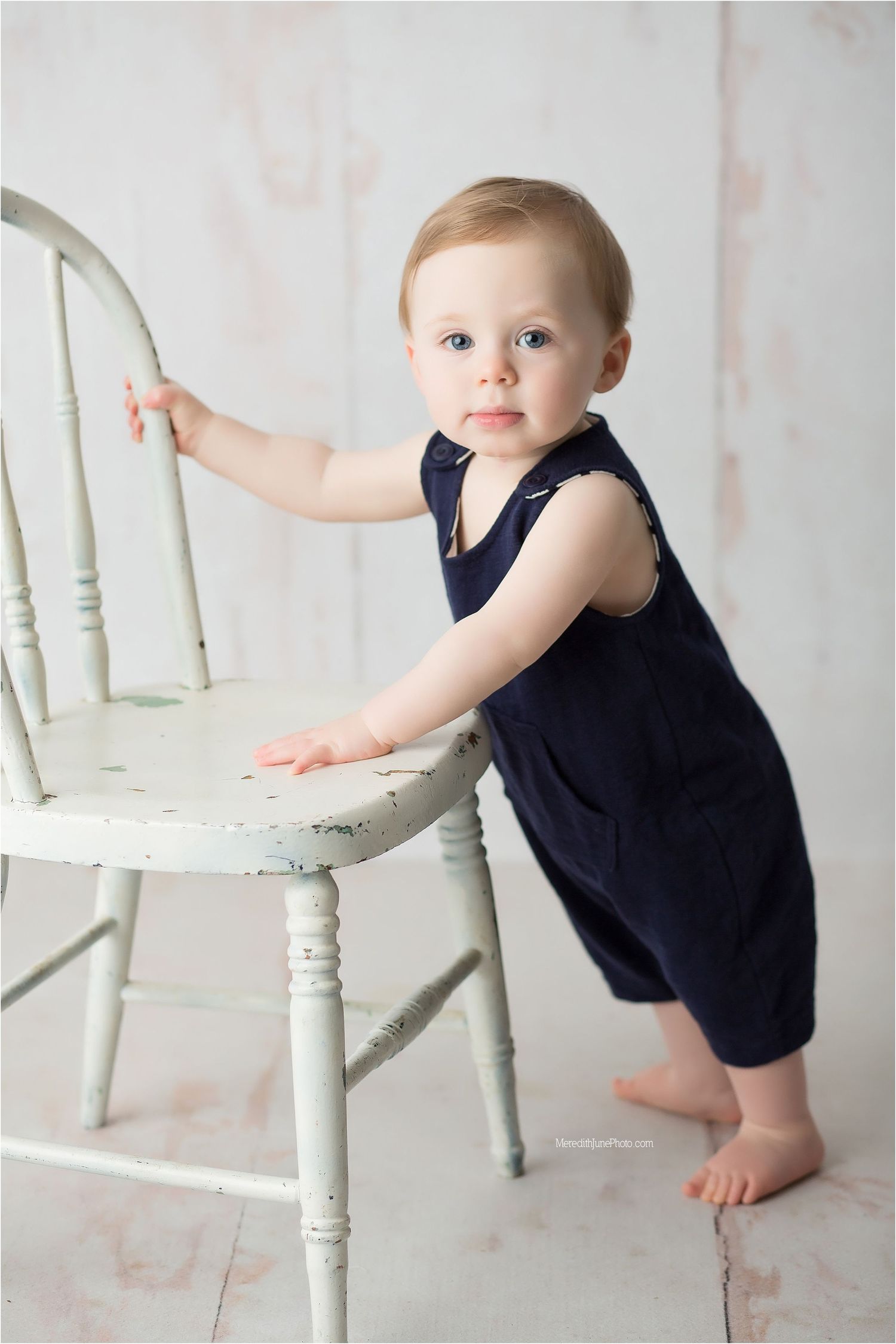 Baby in navy romper practices standing while holding onto white vintage chair in minimalist studio setting.