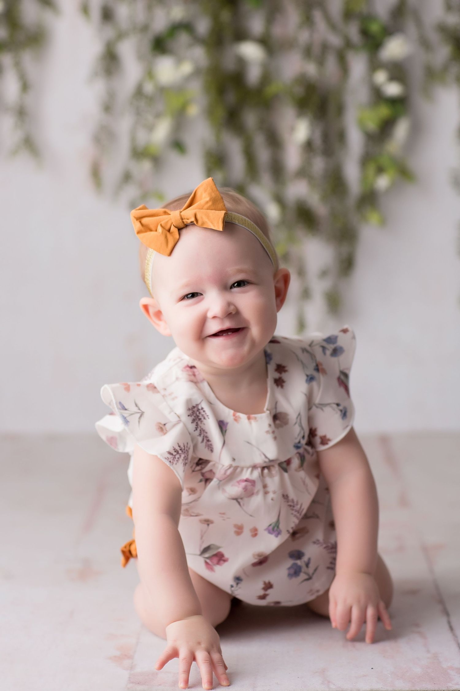 Baby in floral romper with orange bow headband smiles while crawling on light backdrop with greenery.