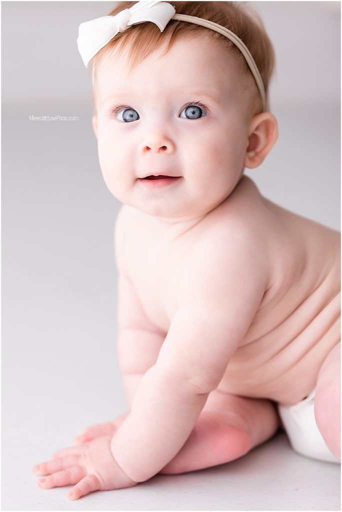 Sweet baby lying on stomach wearing white headband against light gray background in studio setting.