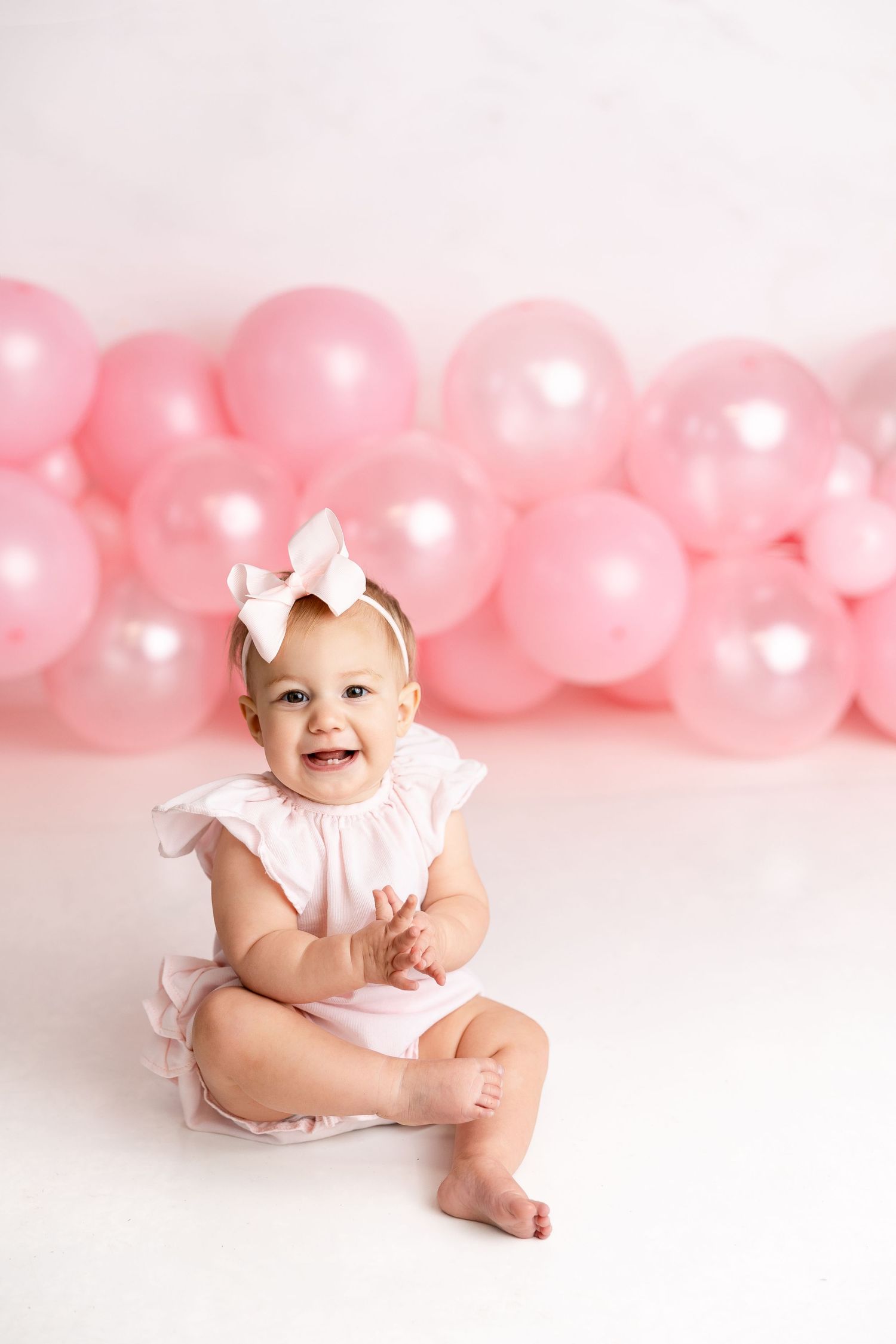 Baby in pink outfit sits on white floor with pink balloon backdrop for milestone photo session.