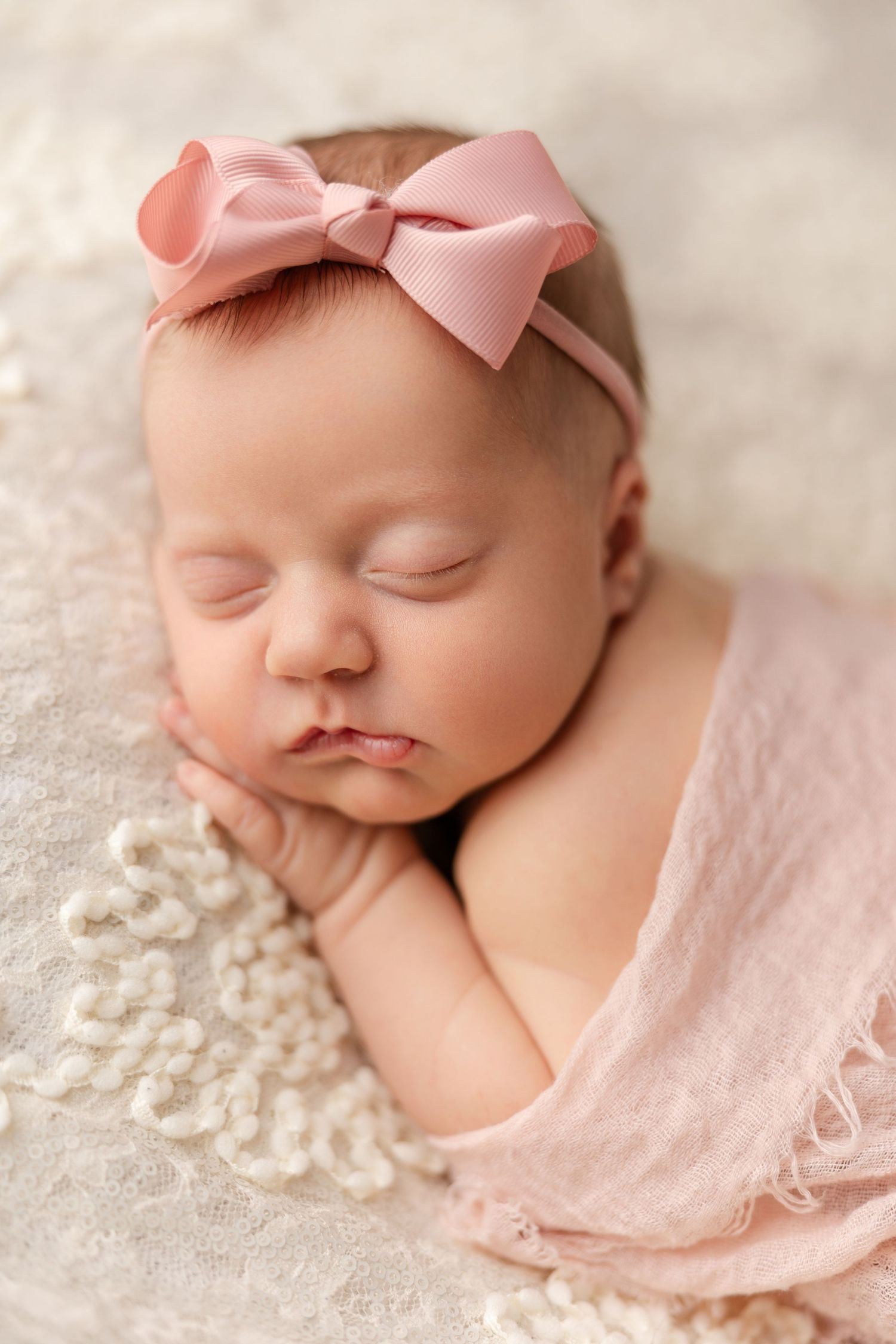 Sleeping newborn in pink wrap and bow headband rests peacefully on delicate lace background.