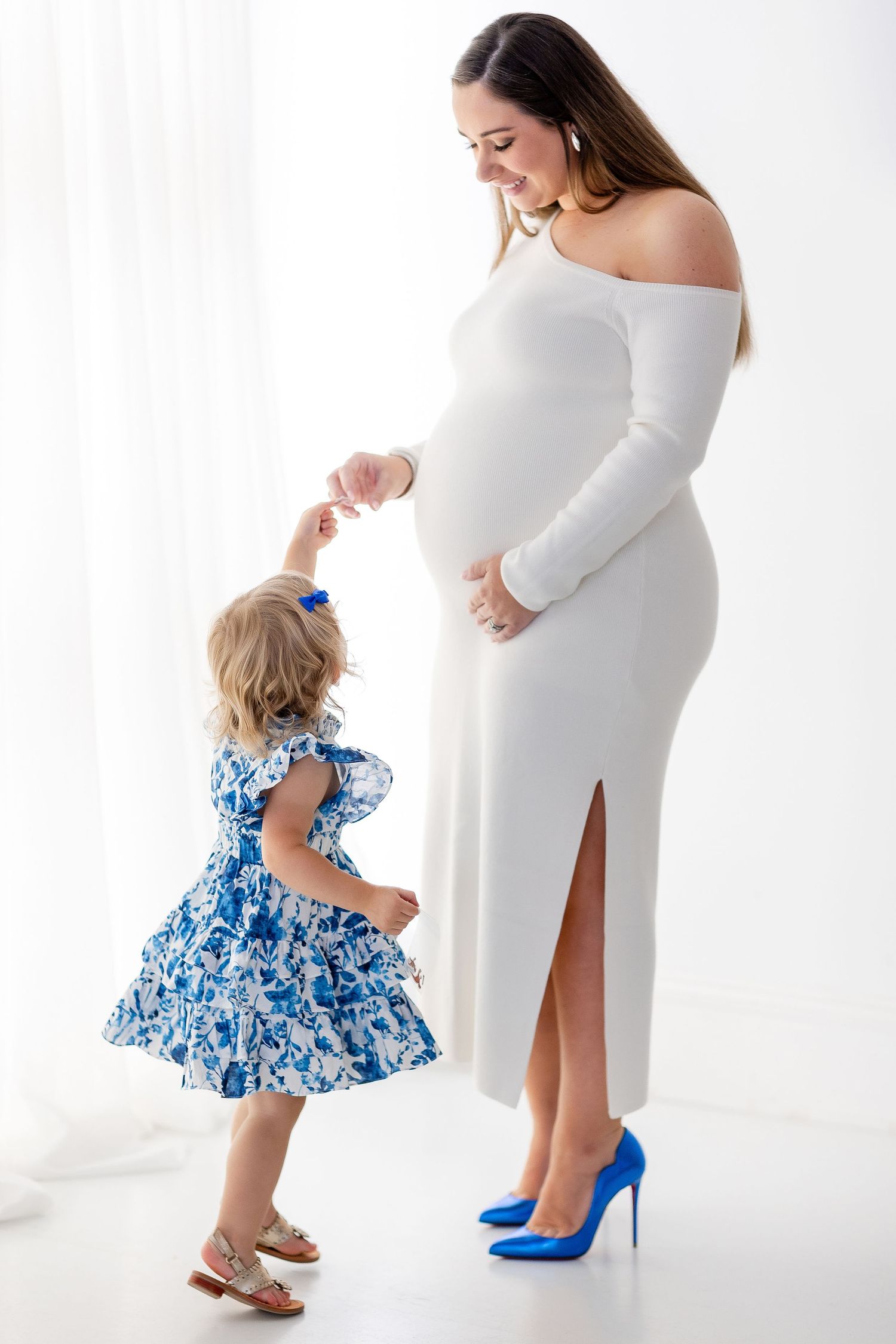 A child in a blue floral dress admires matching blue high heels worn by someone in a white dress against a white backdrop.