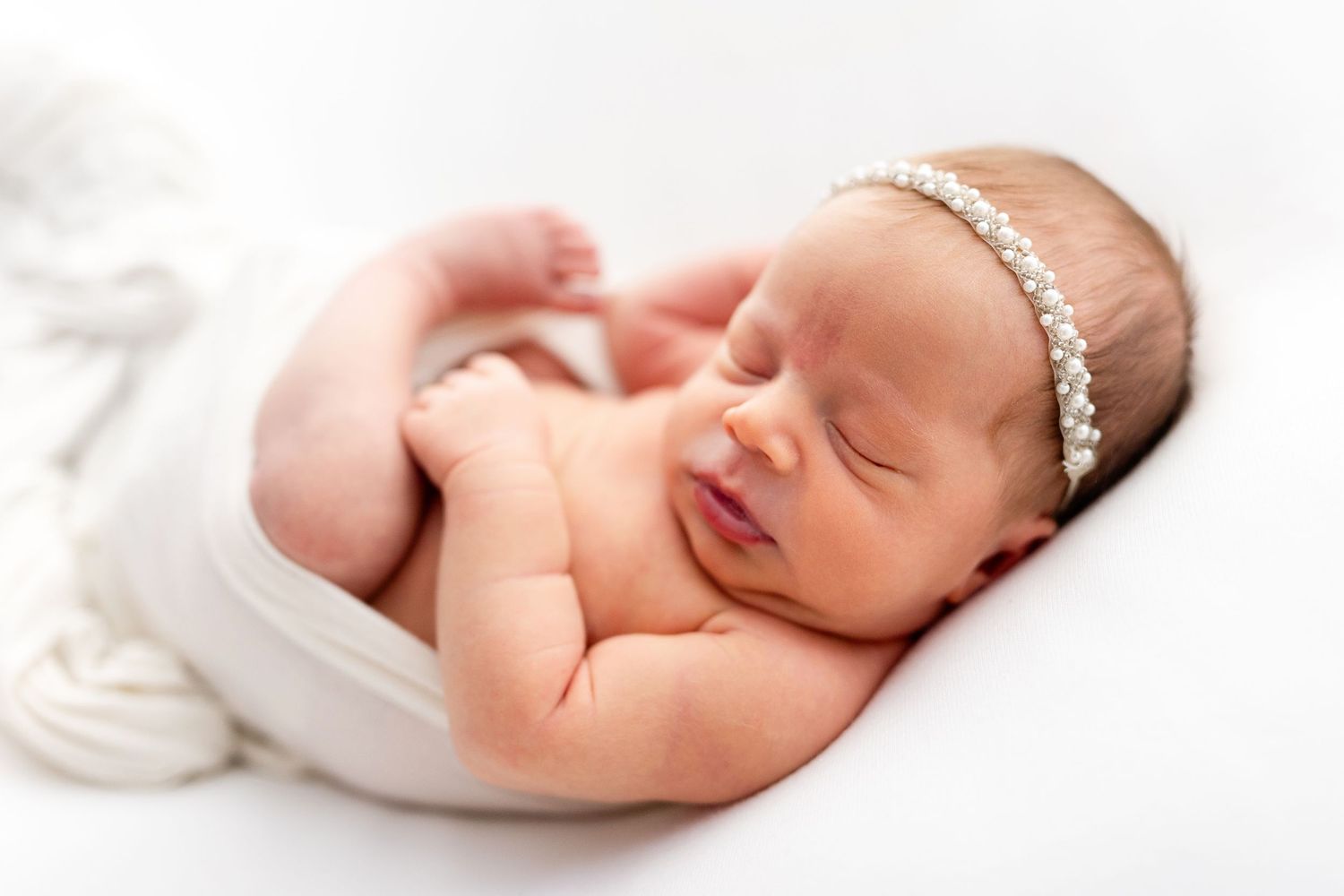 Peaceful sleeping newborn baby wrapped in soft white fabric with delicate headband resting on light background.