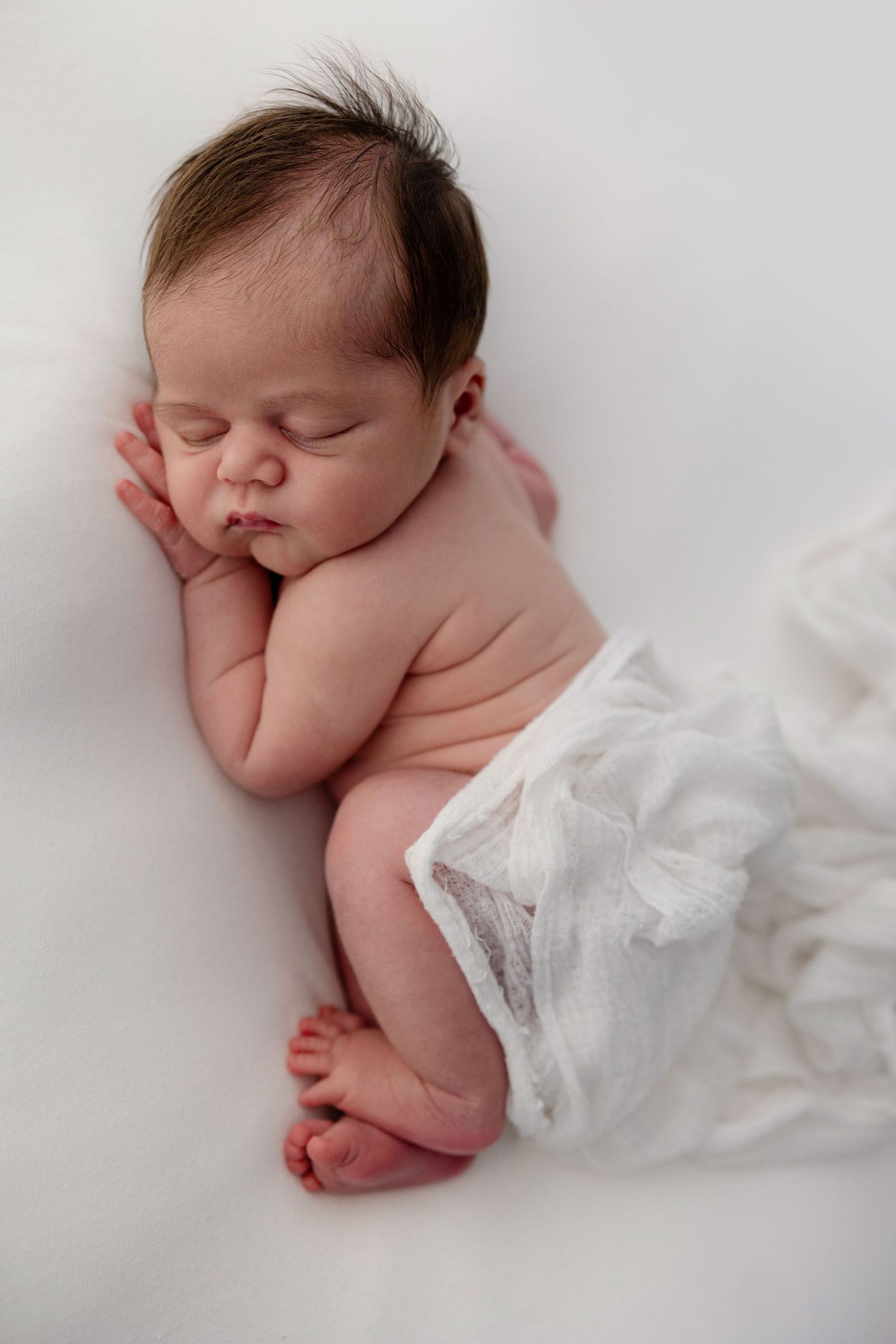 Peaceful newborn baby sleeping on white blanket, dark hair, curled up in fetal position, soft natural lighting.