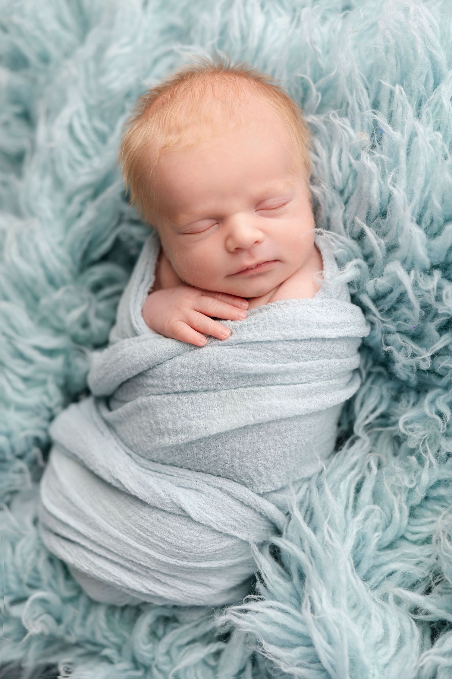 A sleeping newborn baby swaddled in light gray fabric rests peacefully on a soft turquoise textured blanket.